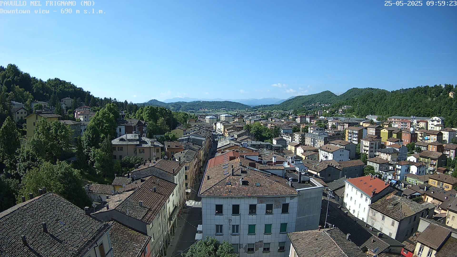 An aerial view shows a town nestled in a valley between green hills under a clear blue sky.
