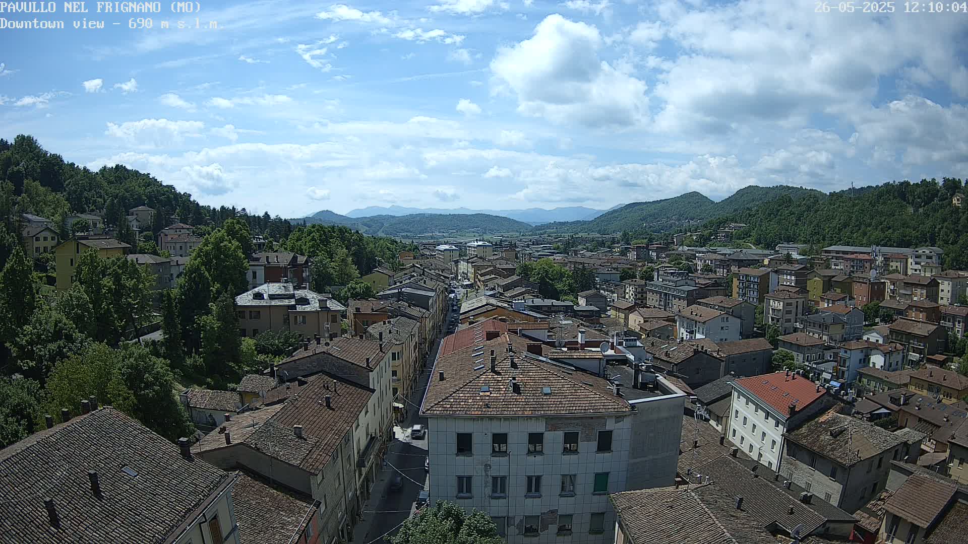 A partly sunny day reveals a view of a town nestled in a valley surrounded by mountains, with numerous buildings and rooftops visible from a high vantage point.