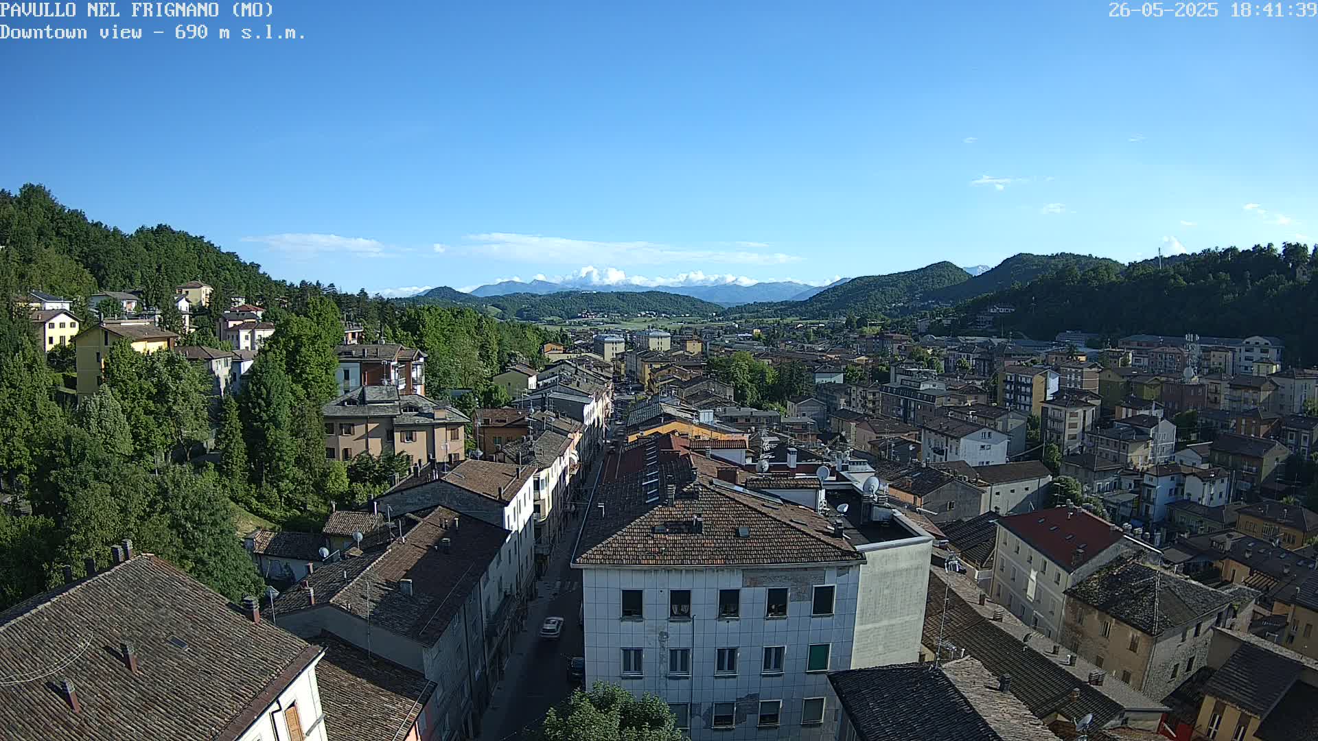 A panoramic view of a town nestled in a valley, under a clear blue sky with a few clouds, shows many buildings with red-tiled roofs and green hills in the background.