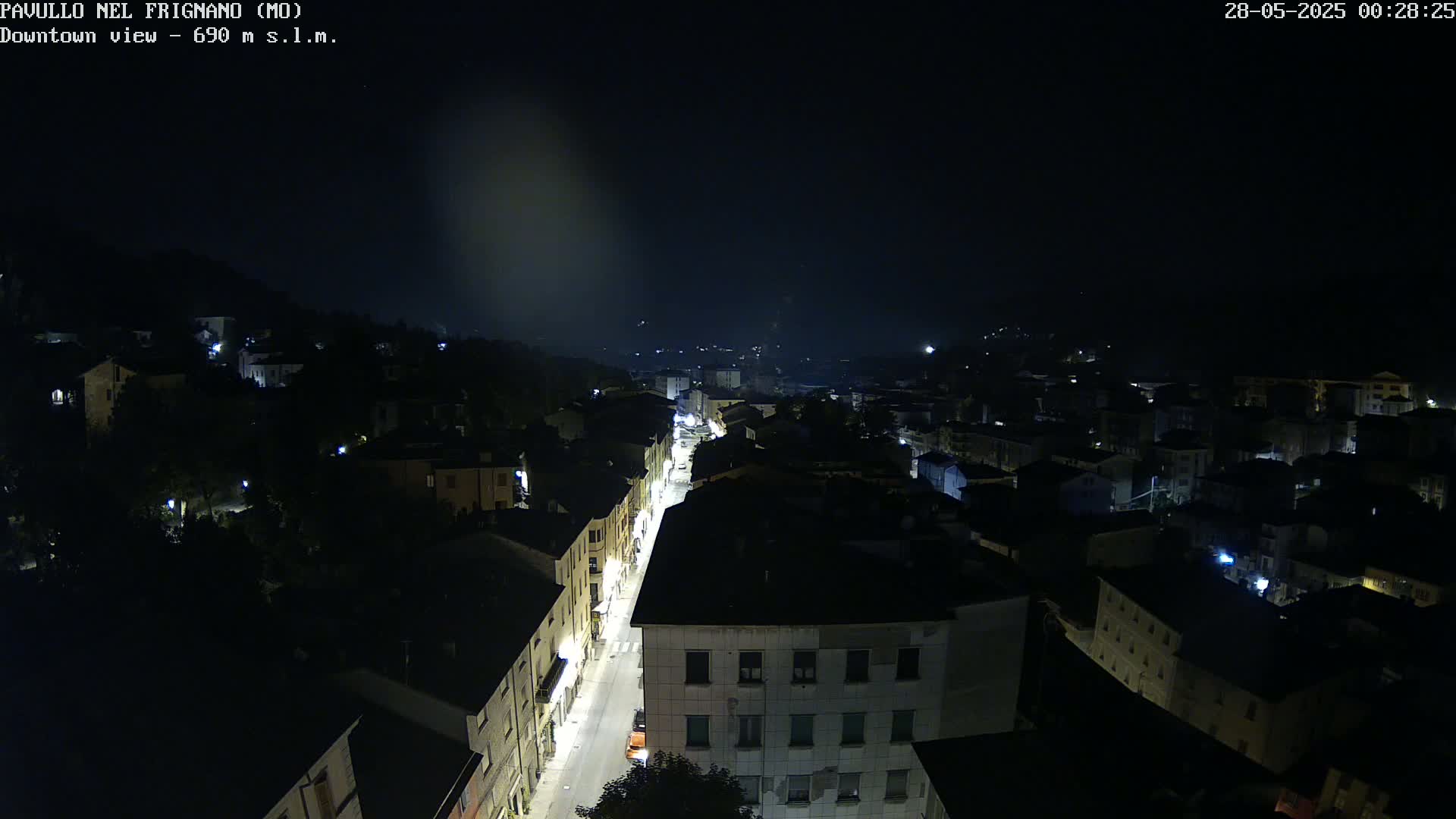 A nighttime aerial view of a town shows a brightly lit street cutting through dark buildings under a hazy, dark sky.
