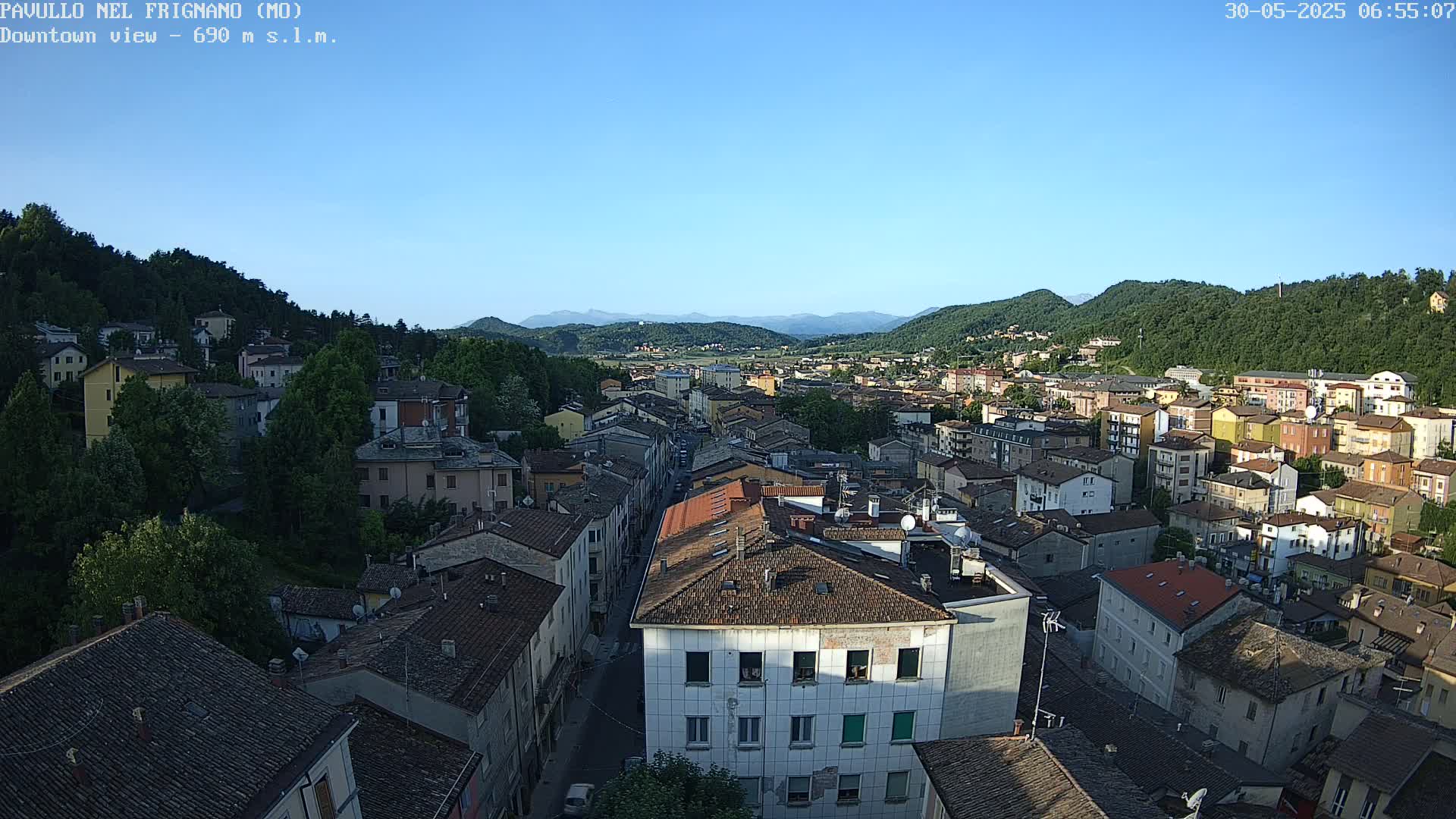 A clear, sunny day reveals a sprawling town nestled in a valley between verdant hills and distant mountains.