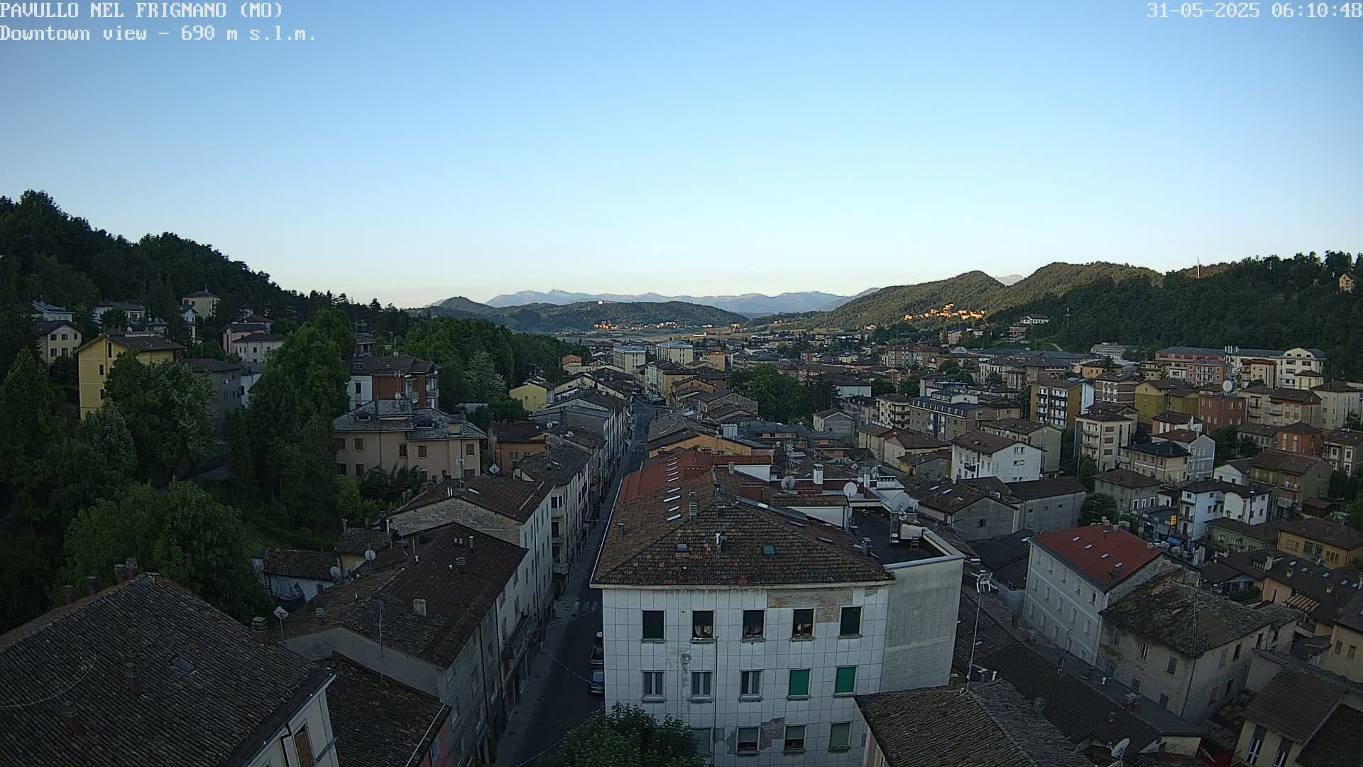 An aerial view of a town nestled in a valley, under a clear blue sky, shows buildings with red-tiled roofs and green hills in the background.