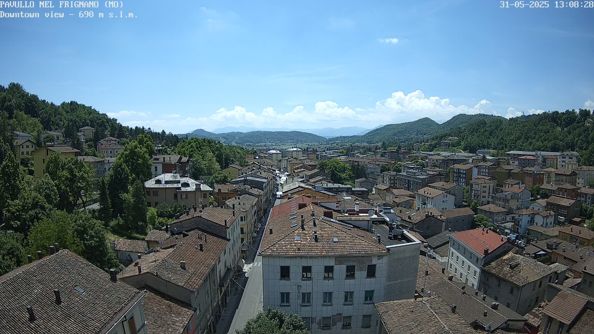 A sunny day reveals a sprawling town nestled in a valley, surrounded by lush green hills and mountains under a partly cloudy sky.