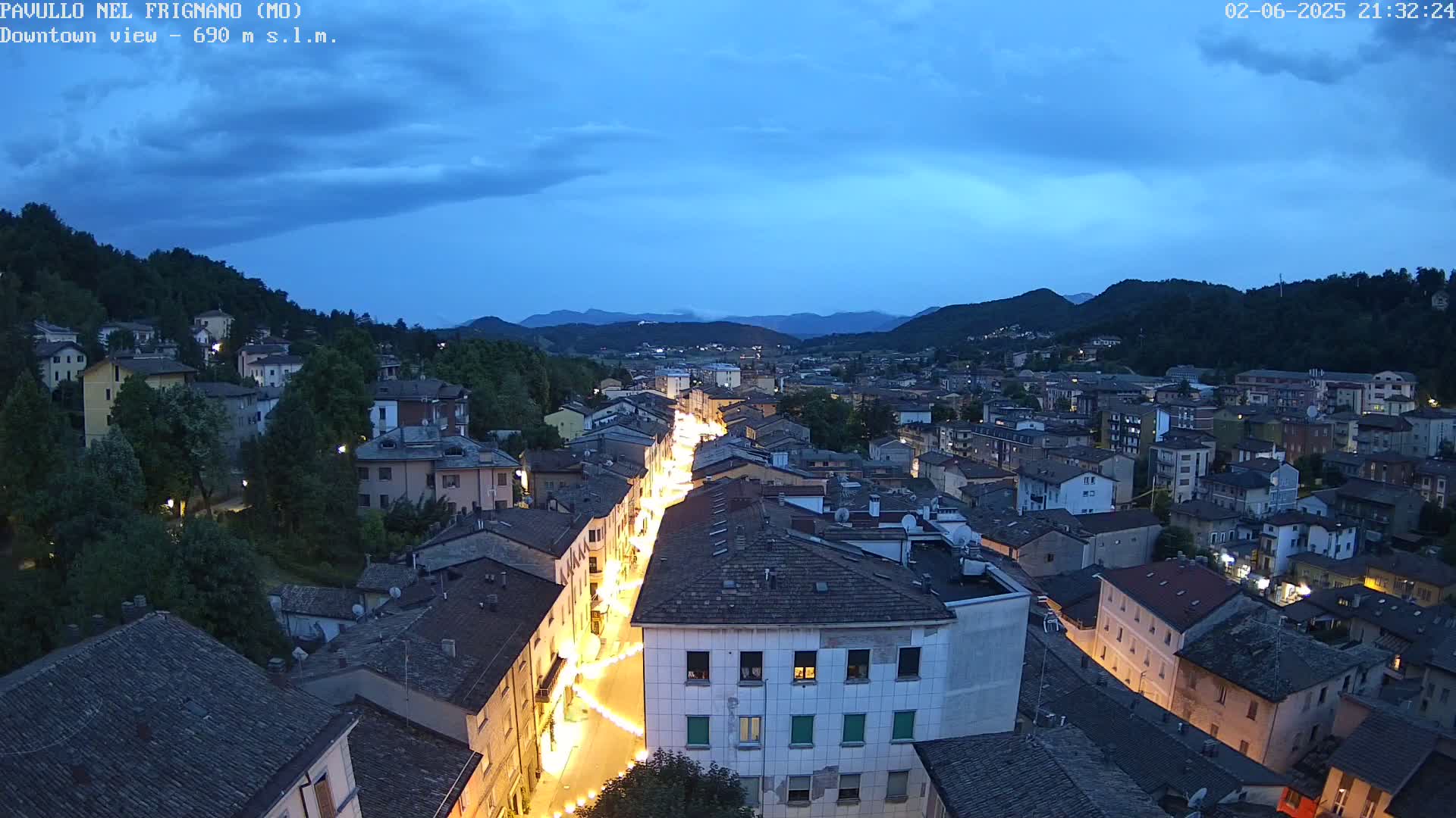 A nighttime, high-angle view of a town nestled in a valley under a twilight sky, with a brightly lit street running through its center.