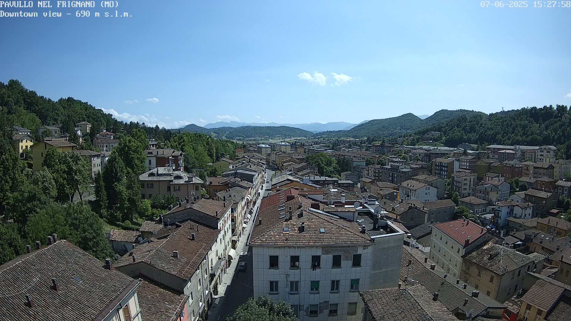An aerial view of a town nestled in a valley, under a mostly sunny sky with a few scattered clouds, shows numerous buildings with red tile roofs surrounded by lush green hills and distant mountains.