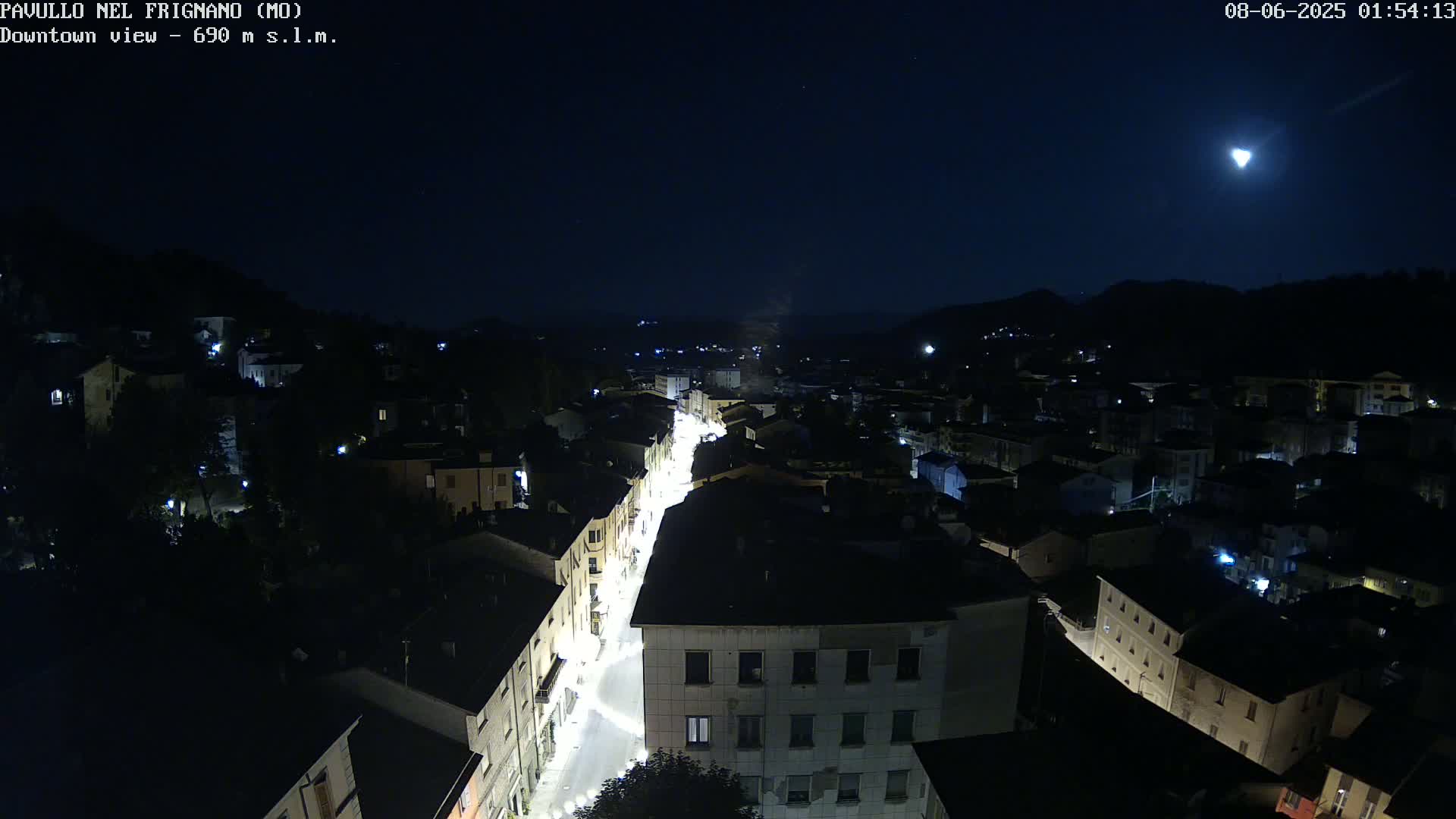 A brightly lit street cuts through a dark, nighttime town nestled in hills under a mostly clear, moonlit sky.