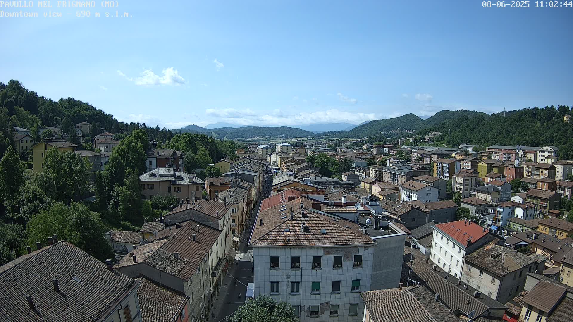 A high-angle view shows a town nestled in a valley under a mostly clear, sunny sky, surrounded by green hills and mountains in the distance.