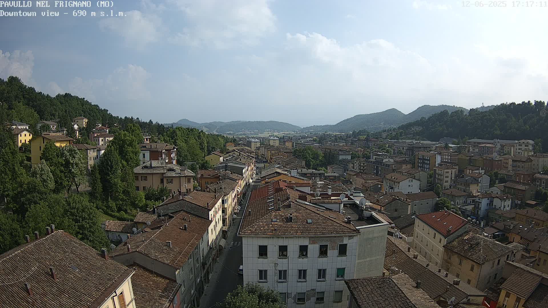 A partly cloudy day reveals a panoramic view of a town nestled in a valley between tree-covered hills.