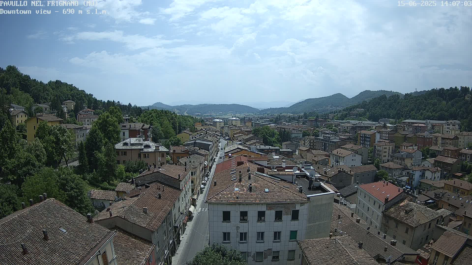 A long street runs through a town nestled in a valley, surrounded by green hills under a partly cloudy sky.