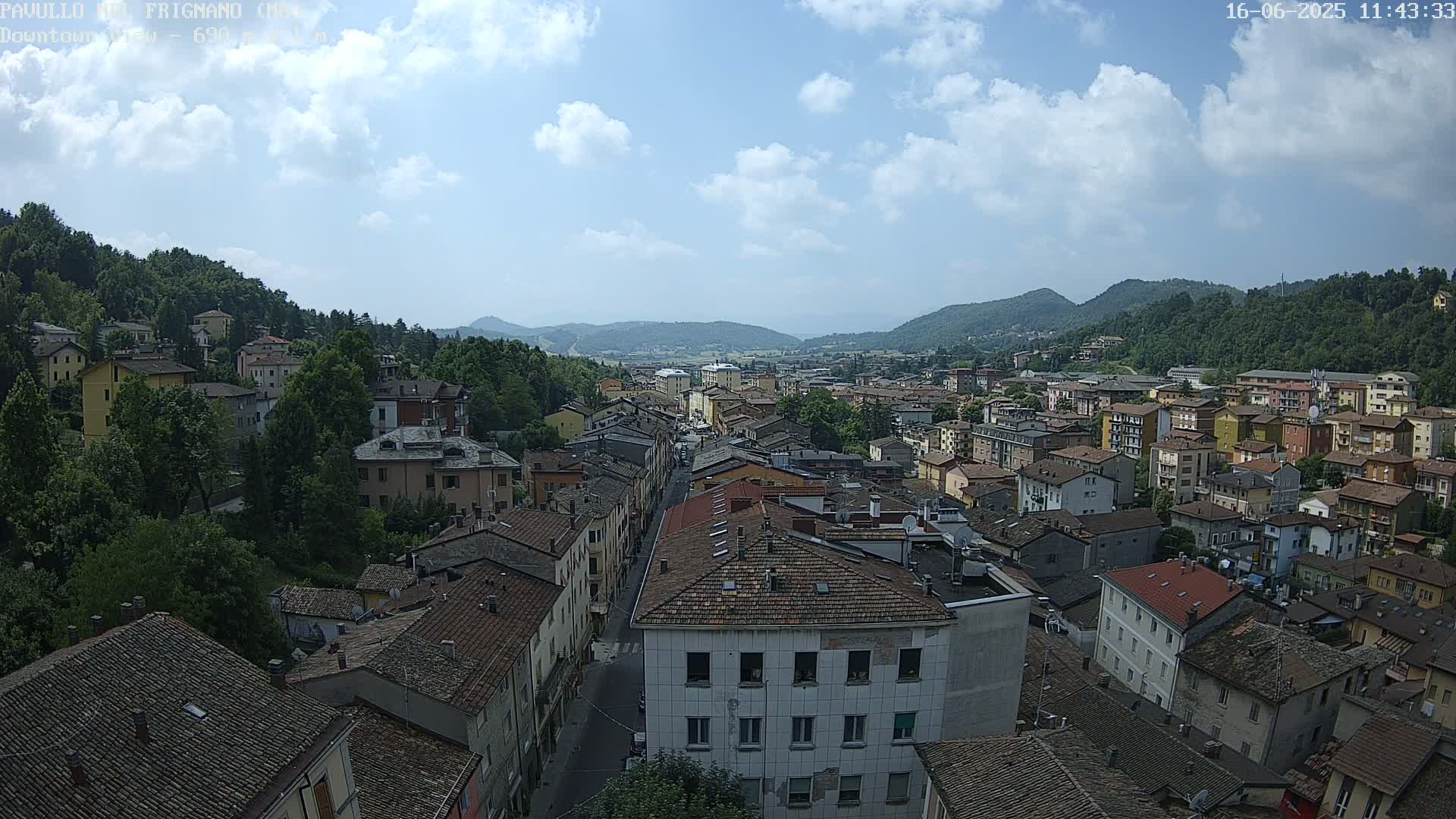 A partly sunny day reveals a sprawling town nestled in a valley between verdant hills, with numerous buildings featuring red-tiled roofs.