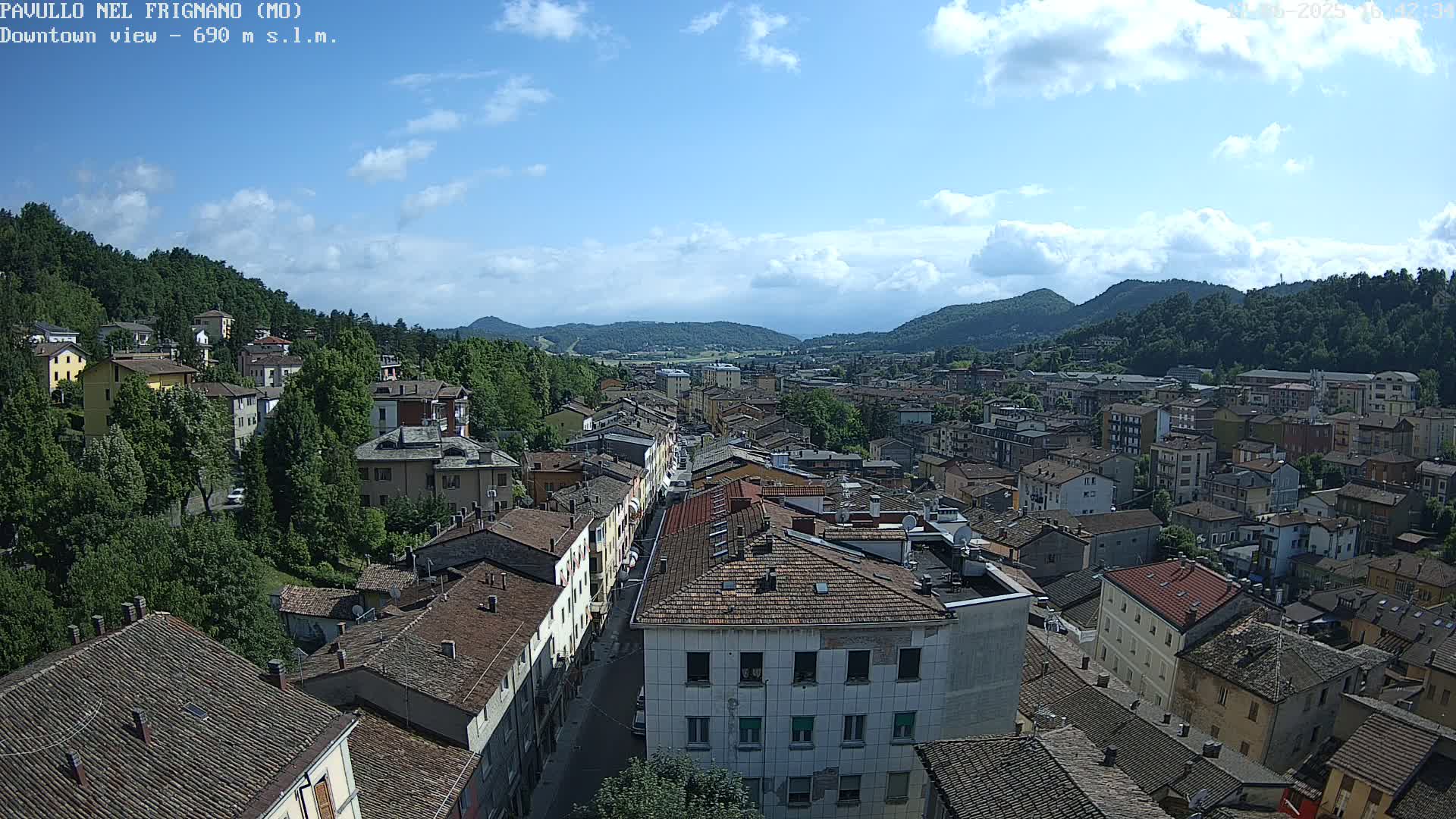 A partly cloudy day reveals an aerial view of a town nestled in a valley surrounded by green hills.