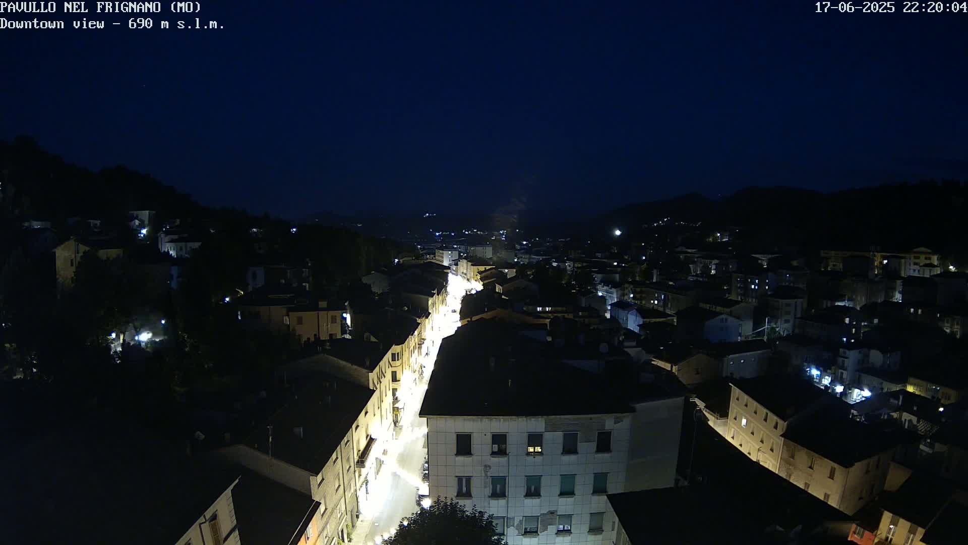 An aerial nighttime view shows a town's illuminated streets and buildings nestled among dark hills under a clear, dark sky.