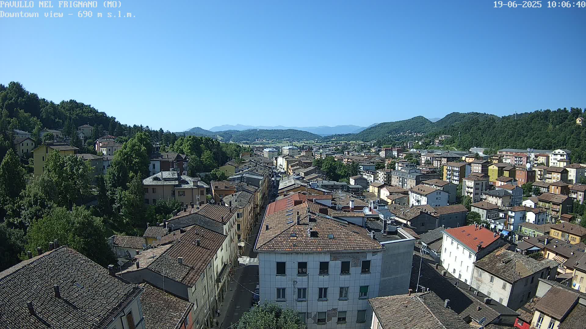 A high-angle, long shot on a clear, sunny day shows a dense collection of buildings in a town nestled in a valley surrounded by green hills and mountains.