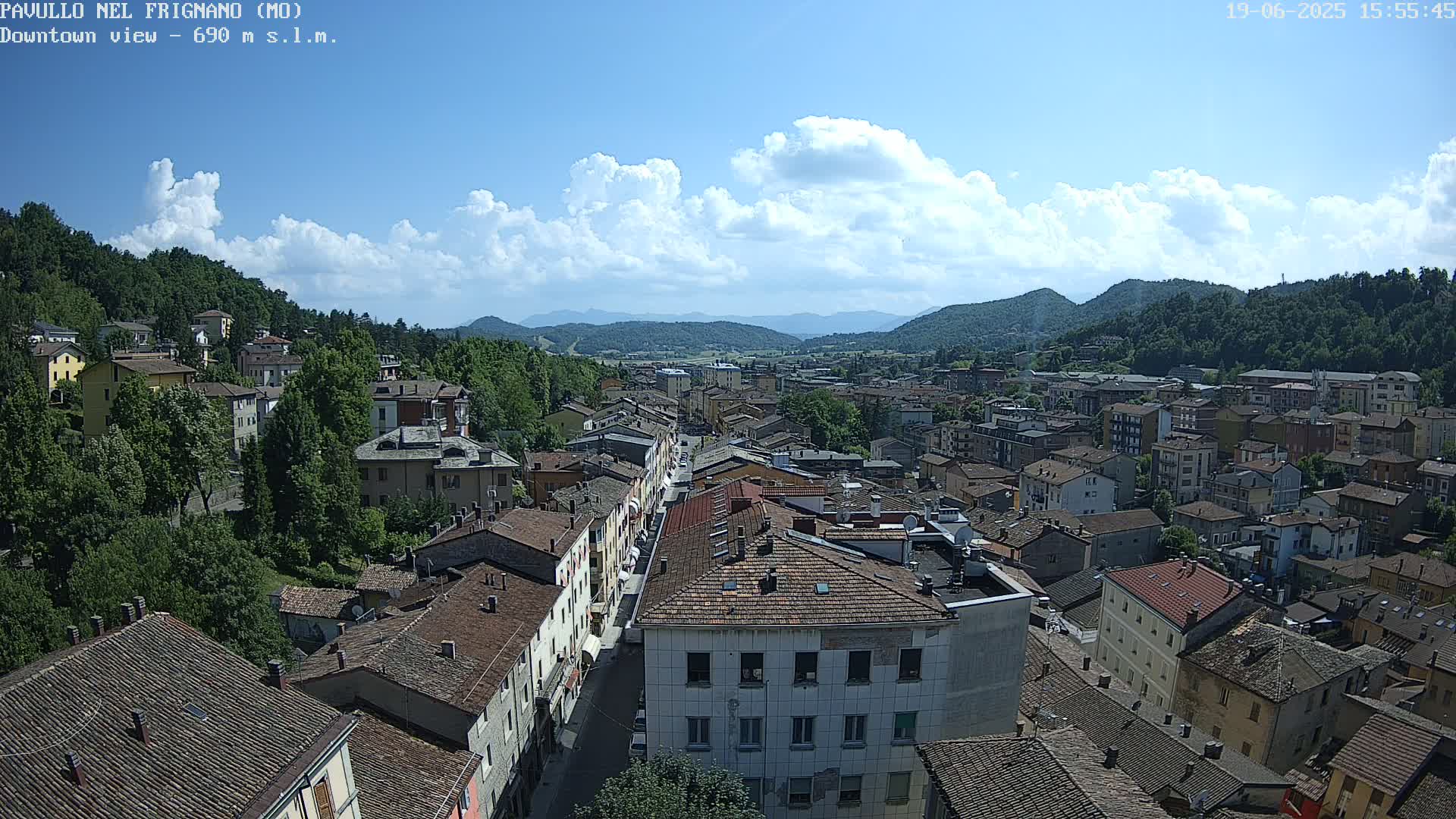A partly cloudy sunny day reveals an aerial view of a town nestled in a valley between green hills.