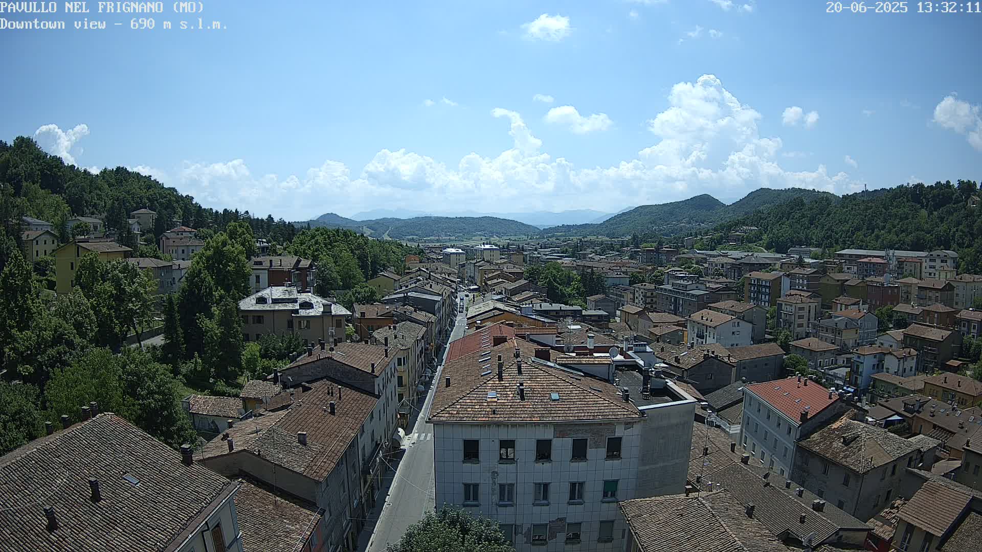 A mostly sunny day reveals a sprawling town nestled in a valley between verdant hills, with a long street running through the center of the town.
