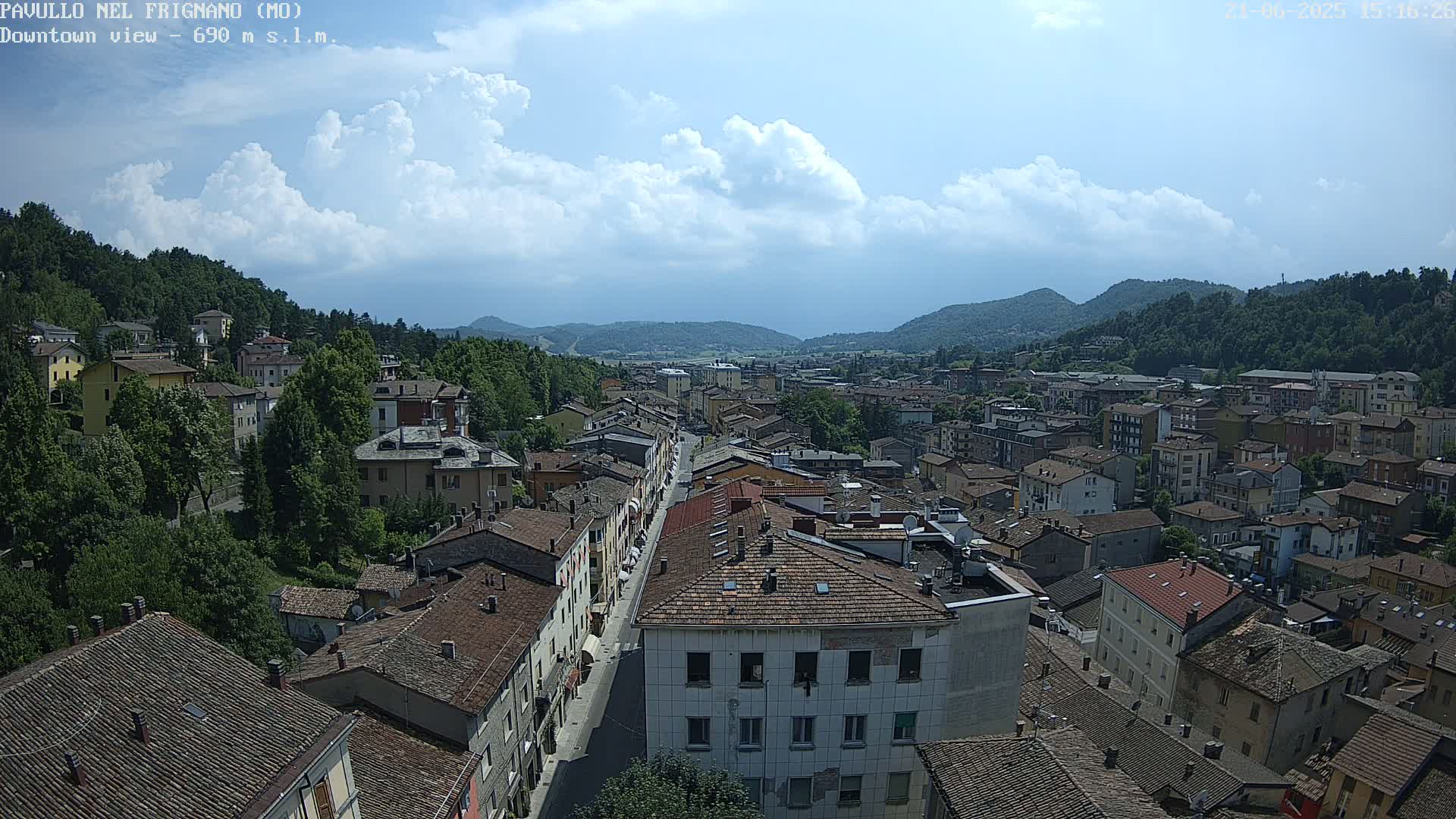 A partly cloudy day reveals a high-angle view of a town nestled in a valley, surrounded by lush green hills.