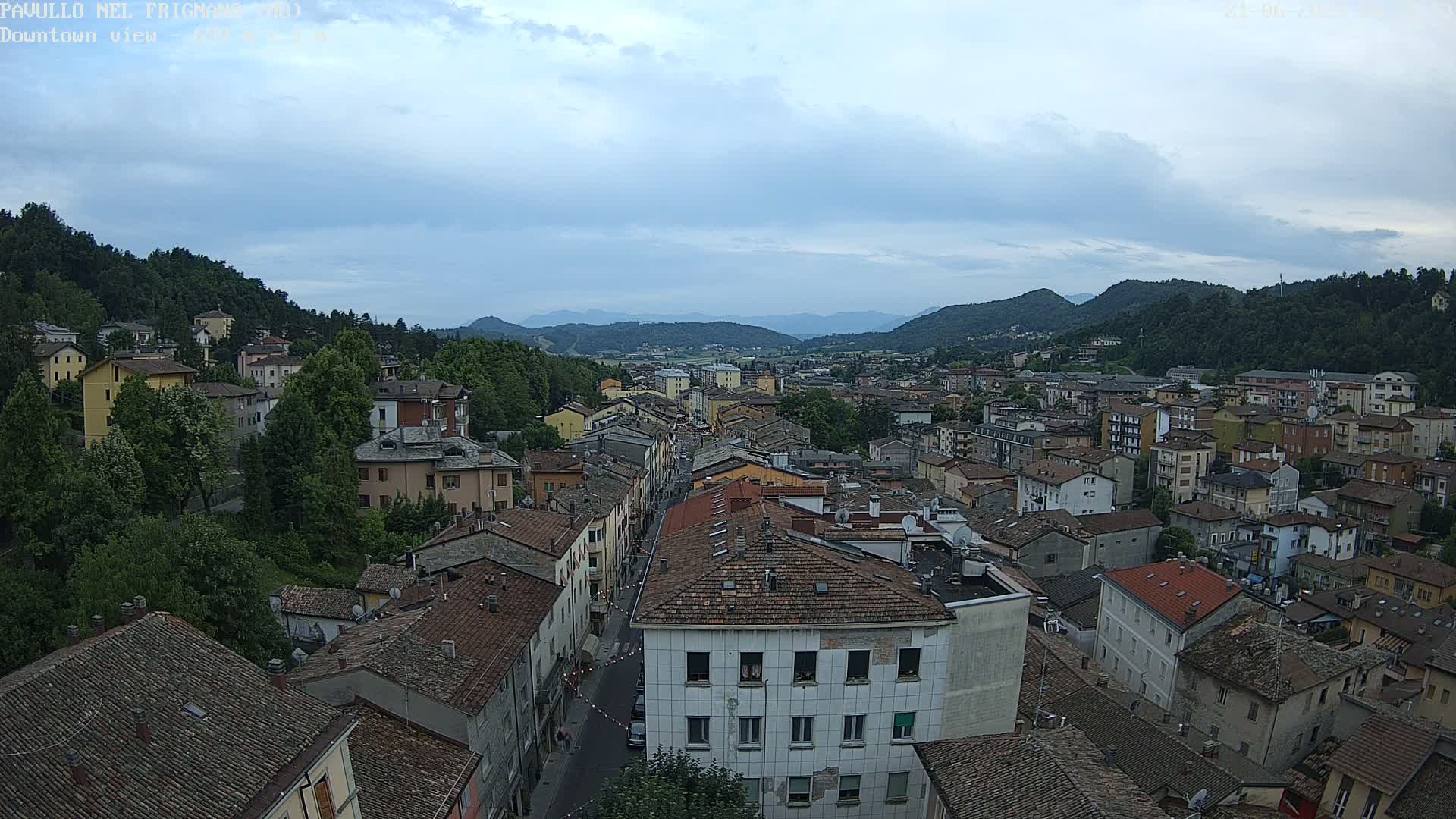 A high-angle view of a town nestled in a valley under a cloudy sky, showing numerous buildings with reddish-brown roofs.