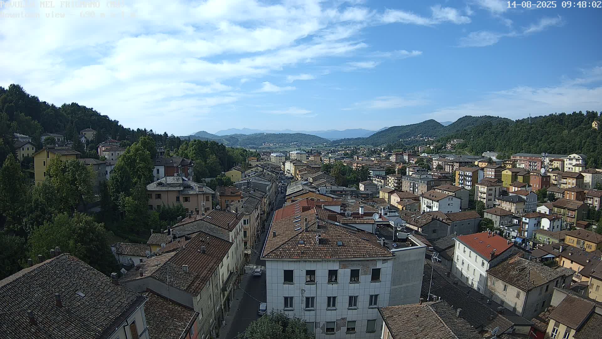 A mostly sunny day reveals a sprawling town nestled in a valley between verdant hills and mountains.