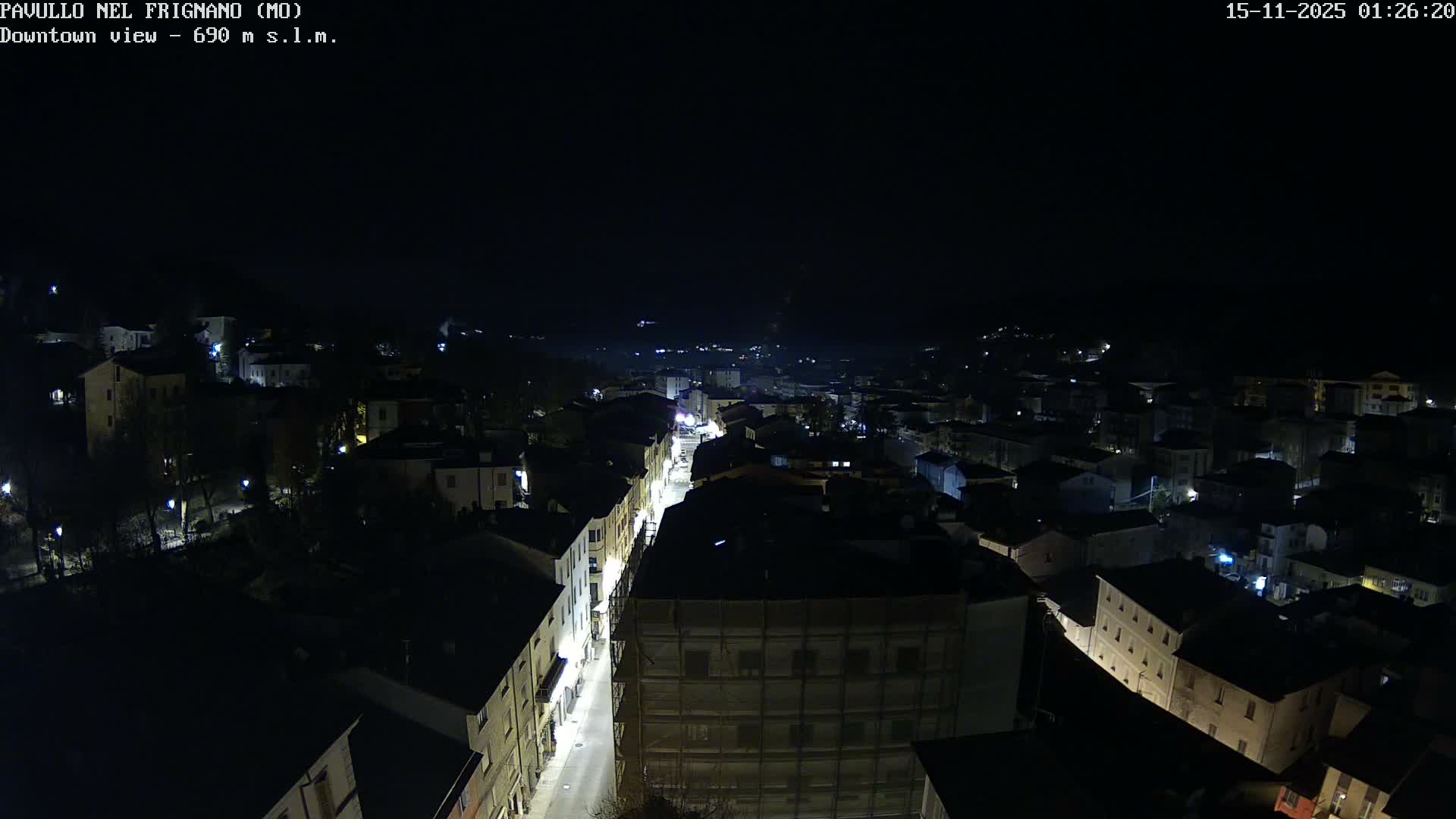 An elevated night view captures a sprawling town illuminated by streetlights and building windows, including one building covered in scaffolding, all beneath a clear, dark sky.