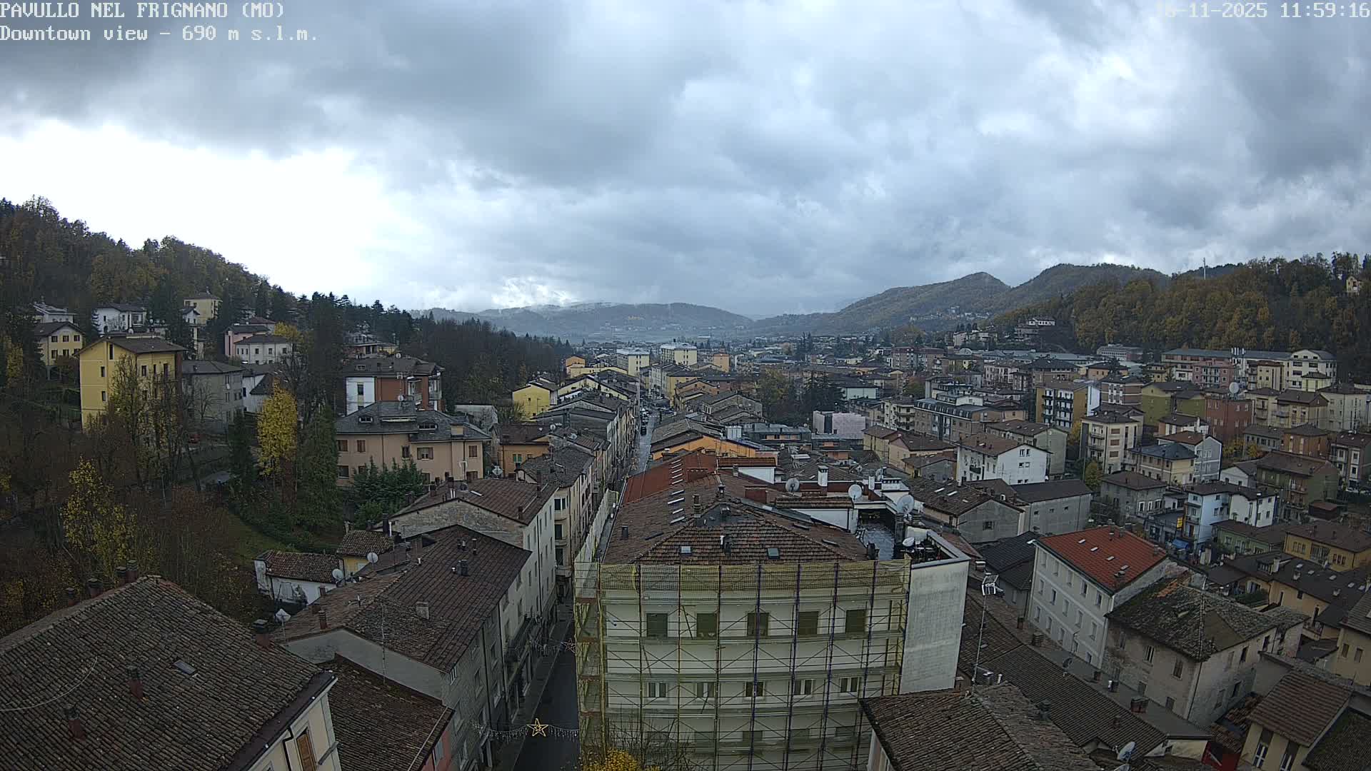 An elevated view captures a dense townscape featuring numerous buildings with tiled roofs stretching through a valley, flanked by tree-covered hills under a heavily overcast sky.
