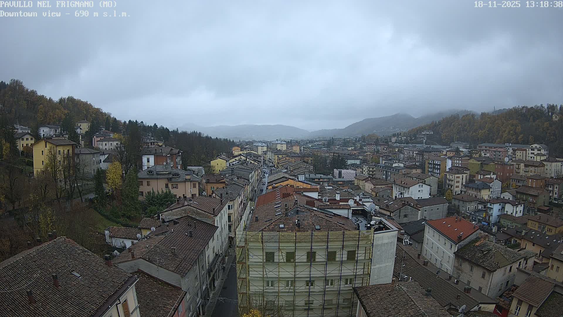 An elevated view captures a dense townscape featuring numerous buildings with tiled roofs stretching through a valley, flanked by tree-covered hills under a heavily overcast sky.