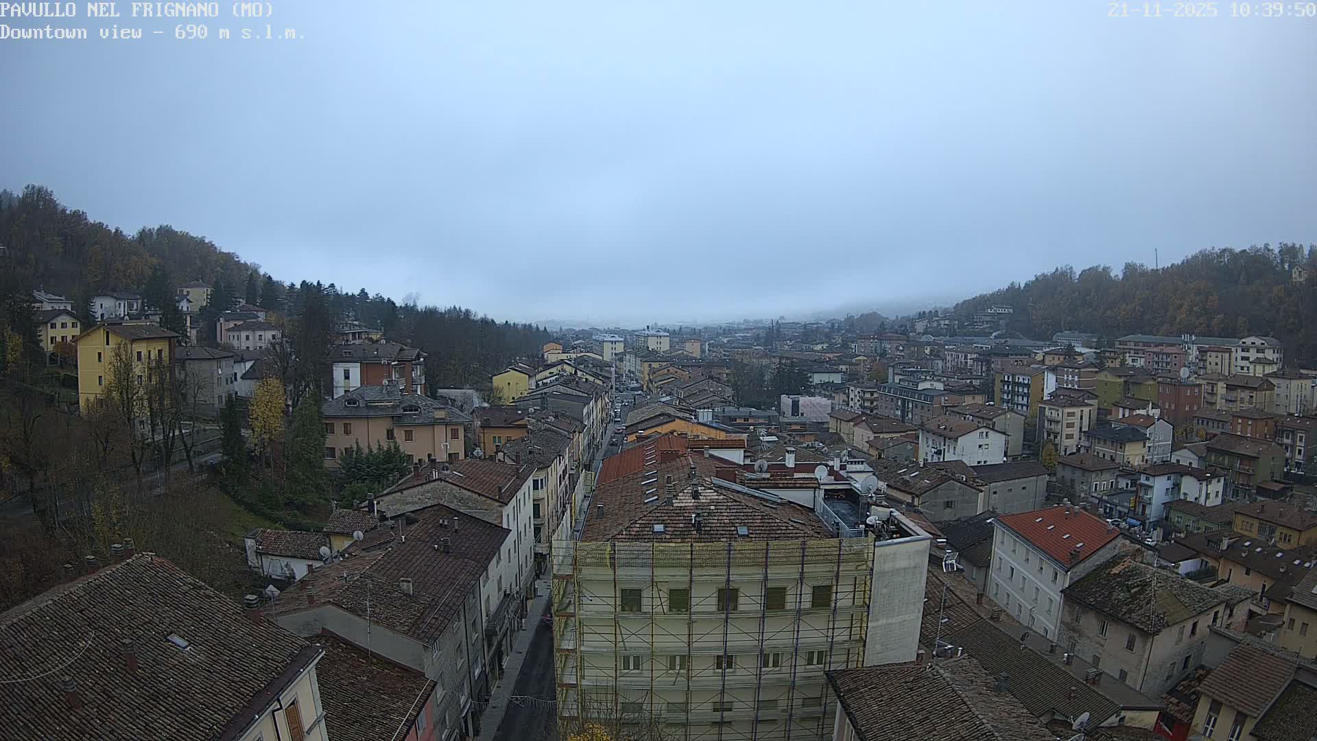 The image shows an expansive aerial view of a densely packed town with traditional buildings nestled in a valley between wooded hills, all under a heavily overcast and hazy sky.