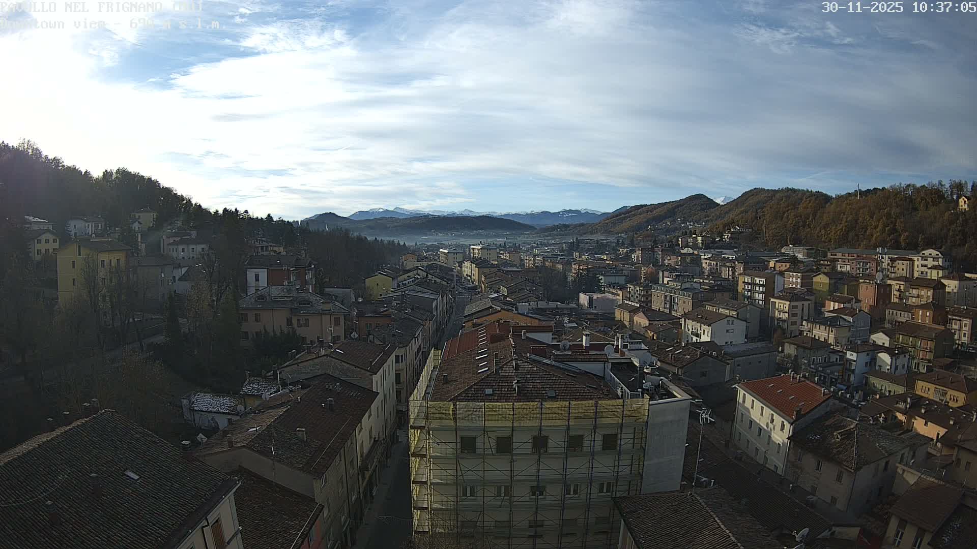 An expansive view reveals a densely packed town with terracotta roofs nestled in a valley between forested hills and distant snow-capped mountains, all under a partly cloudy blue sky.