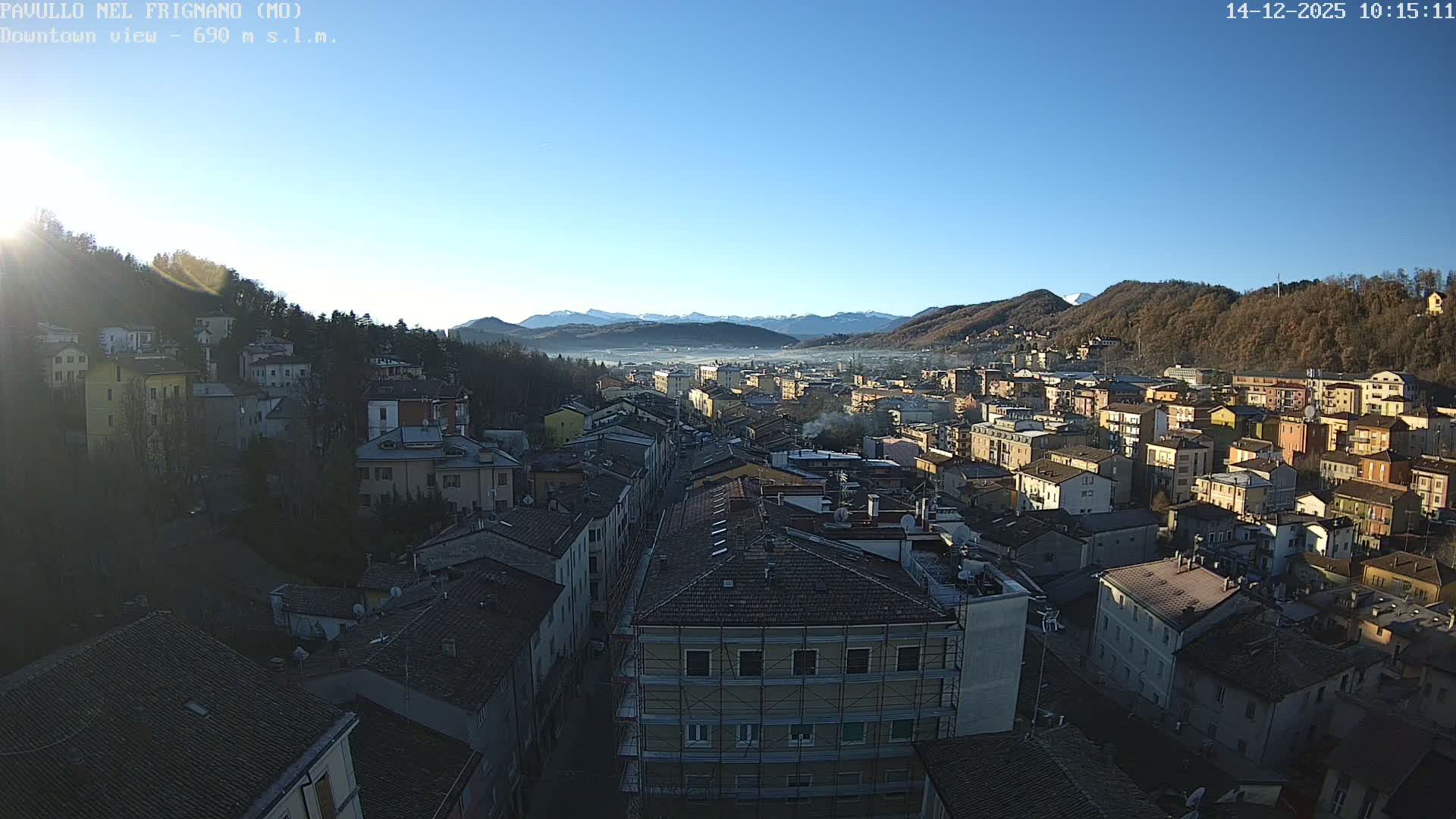 Bright morning sun illuminates a European town sprawling through a valley, flanked by bare-treed hills and distant snow-capped mountains, under a clear blue sky with some fog visible in the far valley.