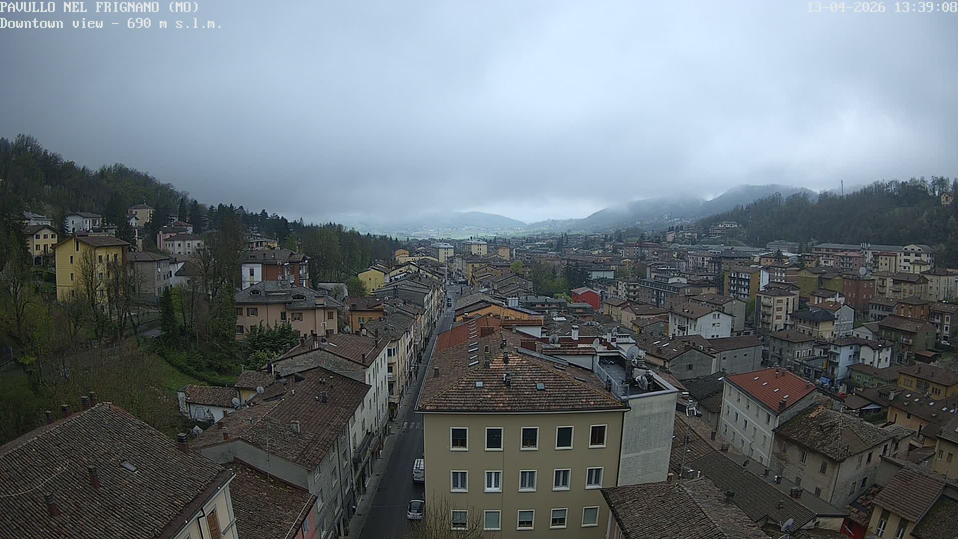 Bright morning sun illuminates a European town sprawling through a valley, flanked by bare-treed hills and distant snow-capped mountains, under a clear blue sky with some fog visible in the far valley.