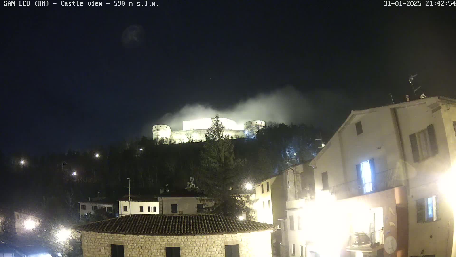 A castle sits atop a hill shrouded in fog at night, overlooking a town with illuminated buildings.