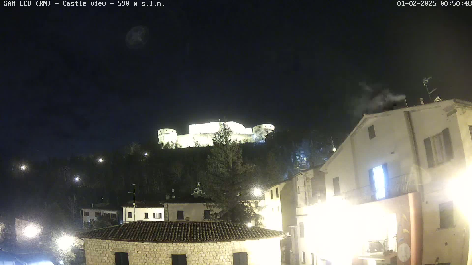 A nighttime view of a hilltop castle illuminated against a dark sky, overlooking a village with several buildings lit by streetlights.