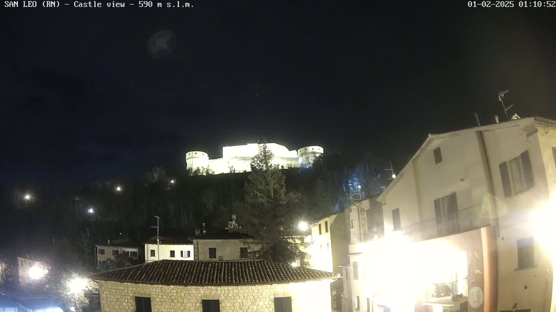 A brightly lit castle sits atop a hill at night, overlooking a town with several buildings illuminated by streetlights under a clear, dark sky.