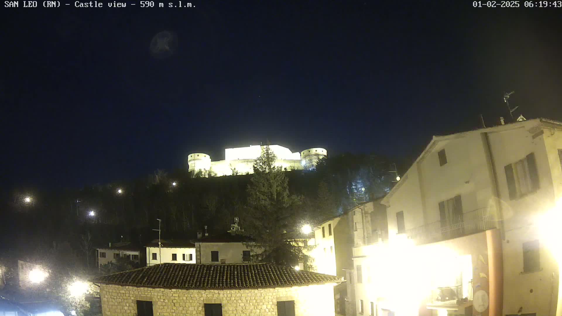 A brightly lit castle sits atop a hill at night, overlooking a town with illuminated buildings.