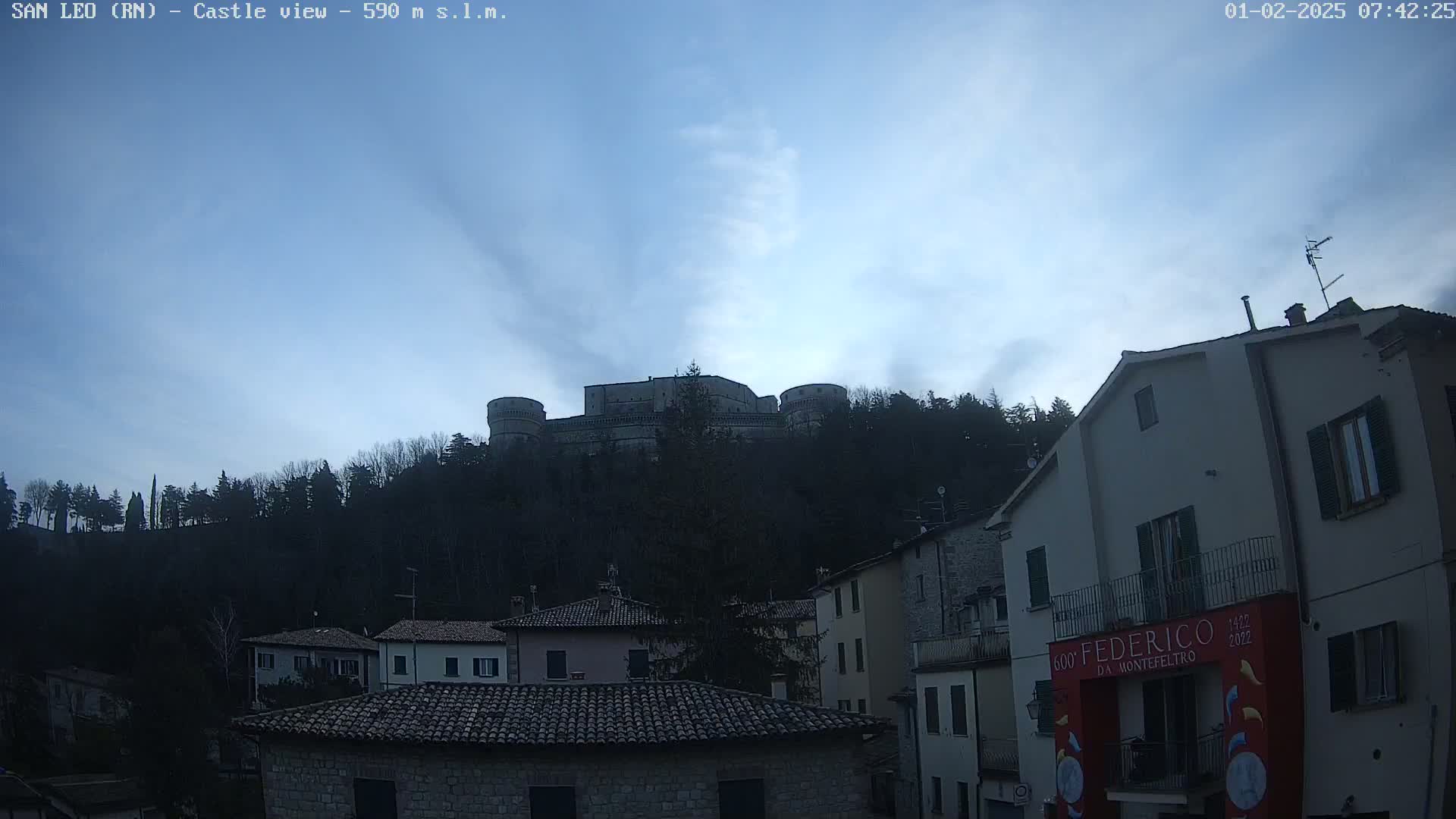 A stone castle sits atop a hill covered in dark trees under a mostly clear, light-blue sky with a few wispy clouds, overlooking a village of stone buildings.
