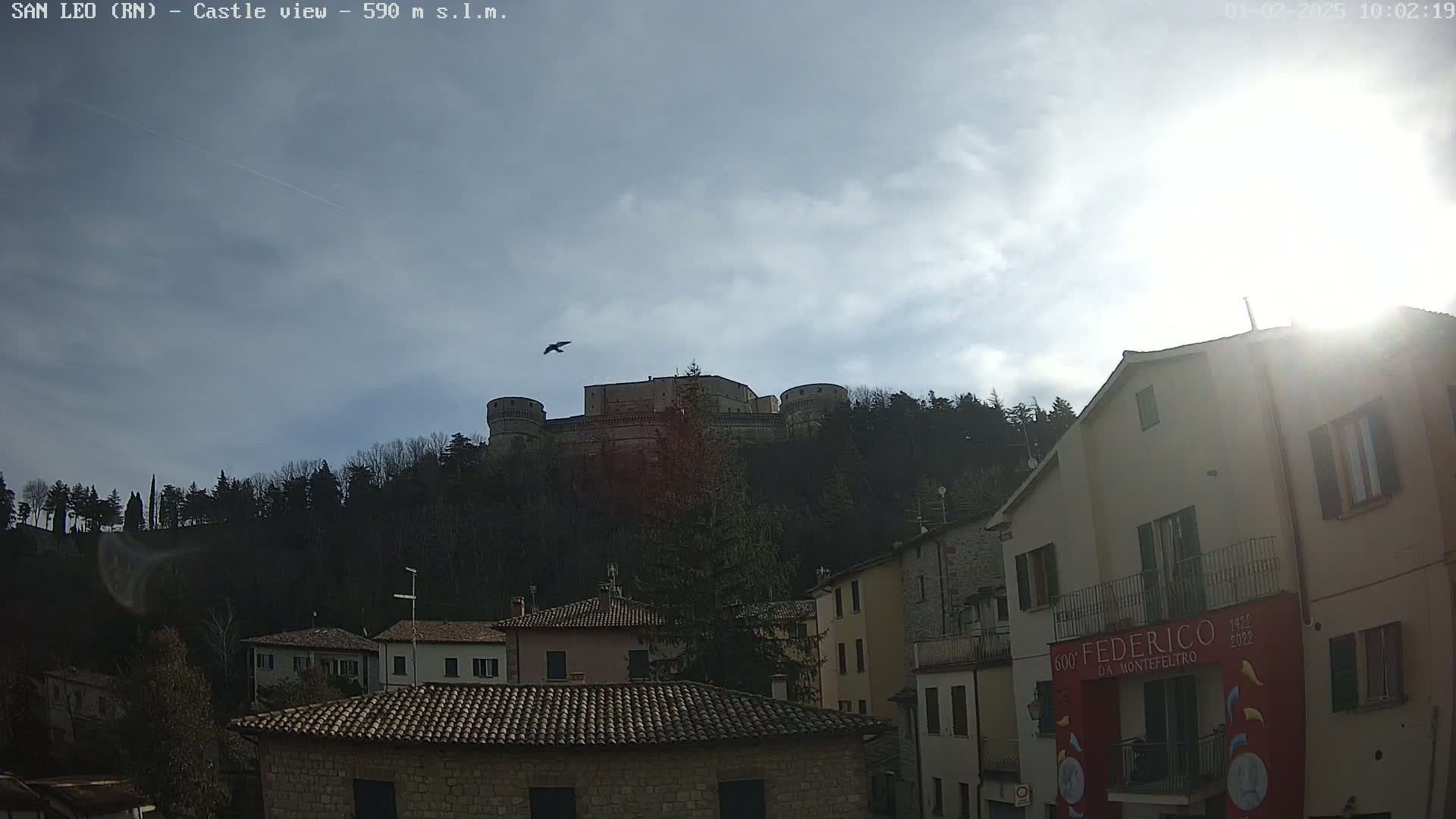 A large stone castle sits atop a hill overlooking a town of buildings with terracotta roofs, under a mostly cloudy sky with a bright sun partially obscured.