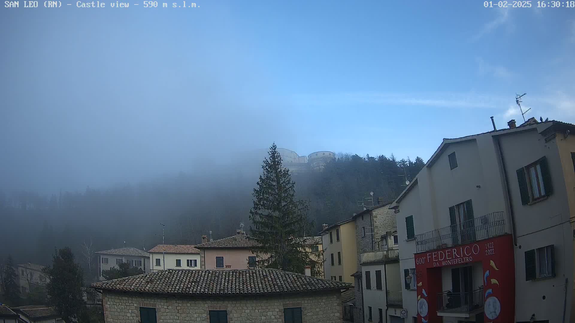 A foggy vista of a town nestled among hills, with a castle visible in the distance under a mostly clear blue sky.