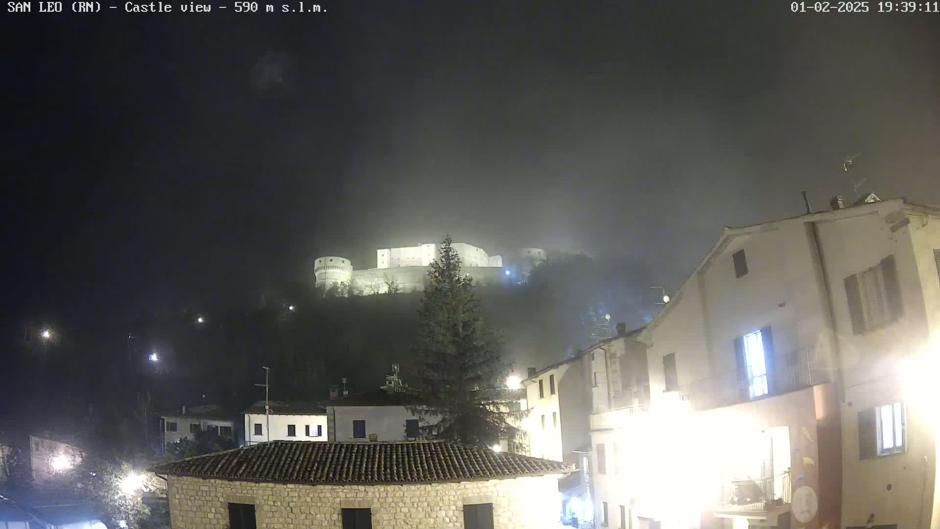 A fog-shrouded castle sits atop a hill overlooking a town at night, illuminated by streetlights.