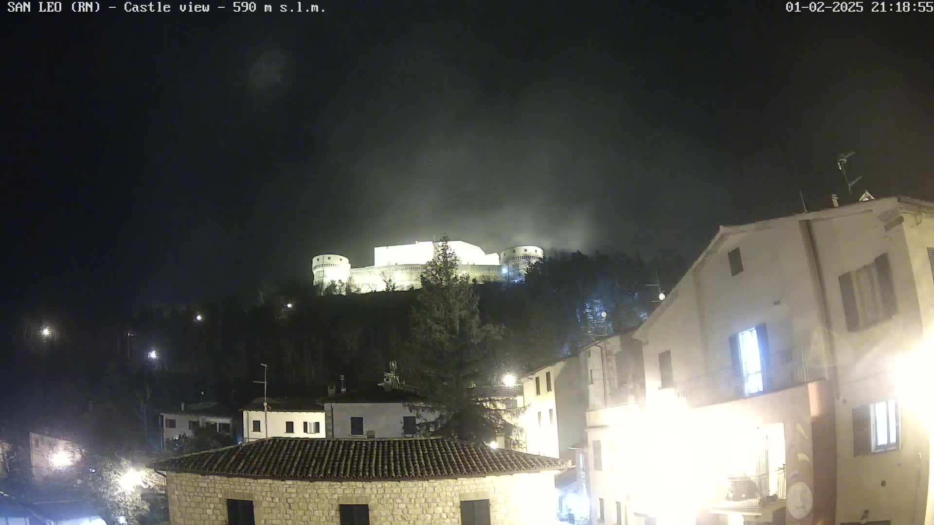 A nighttime view of a hilltop castle illuminated, overlooking a town with buildings and streetlights under a hazy, dark sky.