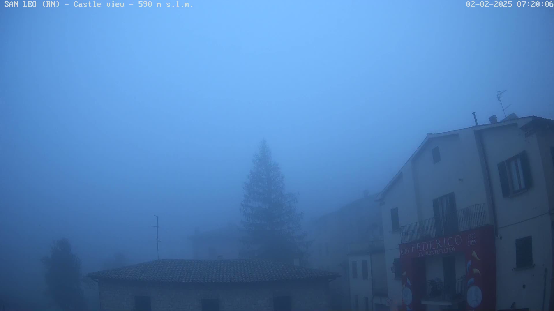 A foggy village scene shows several stone buildings shrouded in a thick blue fog, with a lone evergreen tree visible in the middle ground.