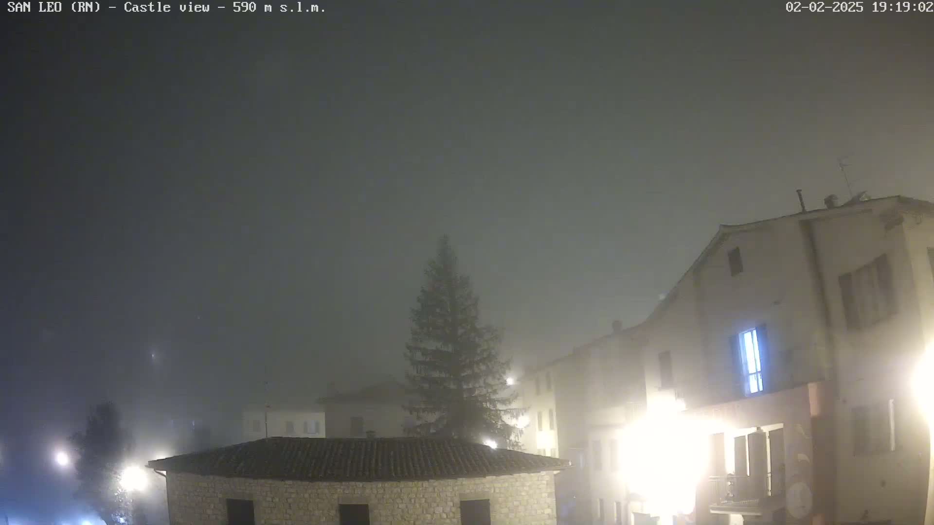 A foggy night scene shows a small town square with stone buildings and a tall evergreen tree, illuminated by streetlights.