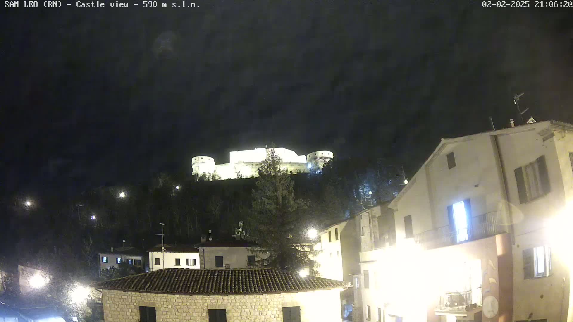 A nighttime view of a hilltop castle illuminated against a dark, clear sky, overlooking a small town with buildings lit by streetlights.