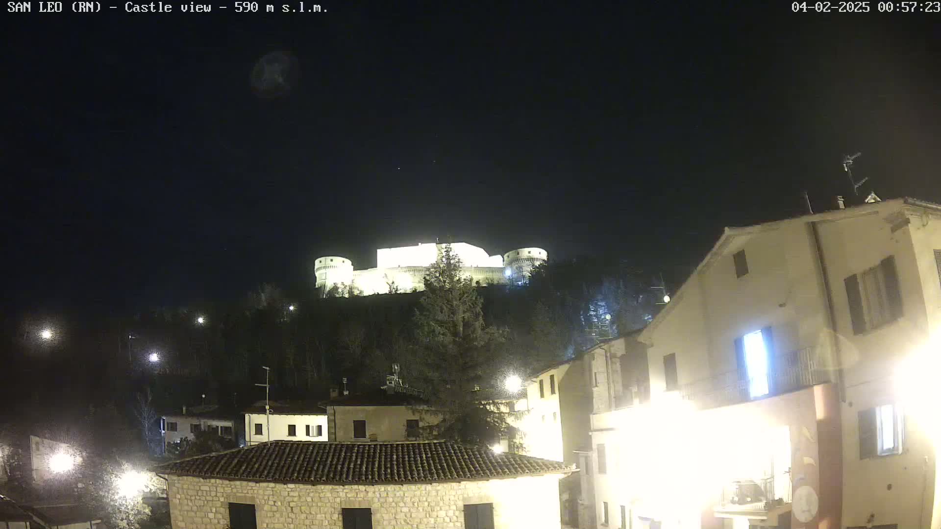 A brightly lit castle sits atop a hill overlooking a village at night under a clear, dark sky.