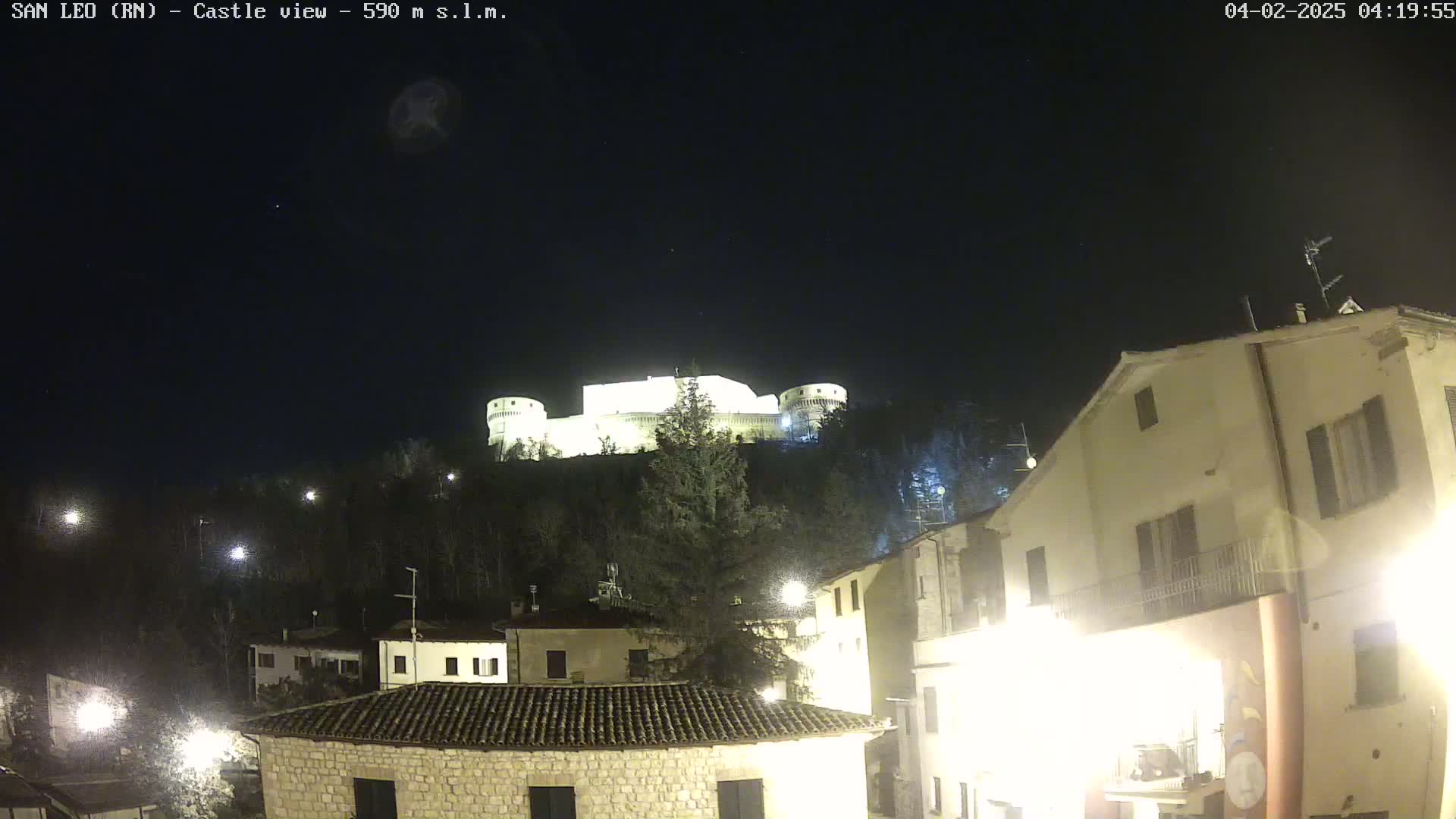 A brightly lit castle sits atop a hill overlooking a small town at night under a clear, dark sky.