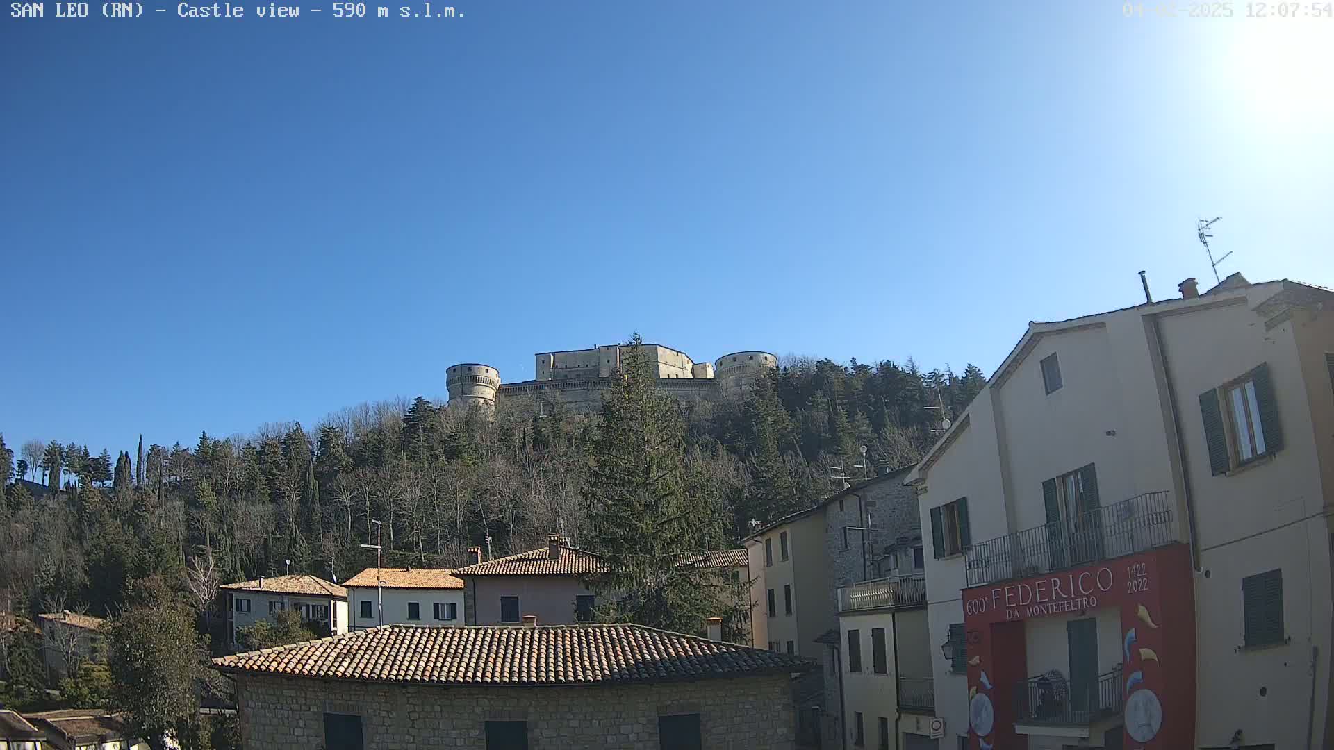 A large stone castle sits atop a hill covered in leafless trees under a clear blue sky, overlooking a town of low stone buildings.