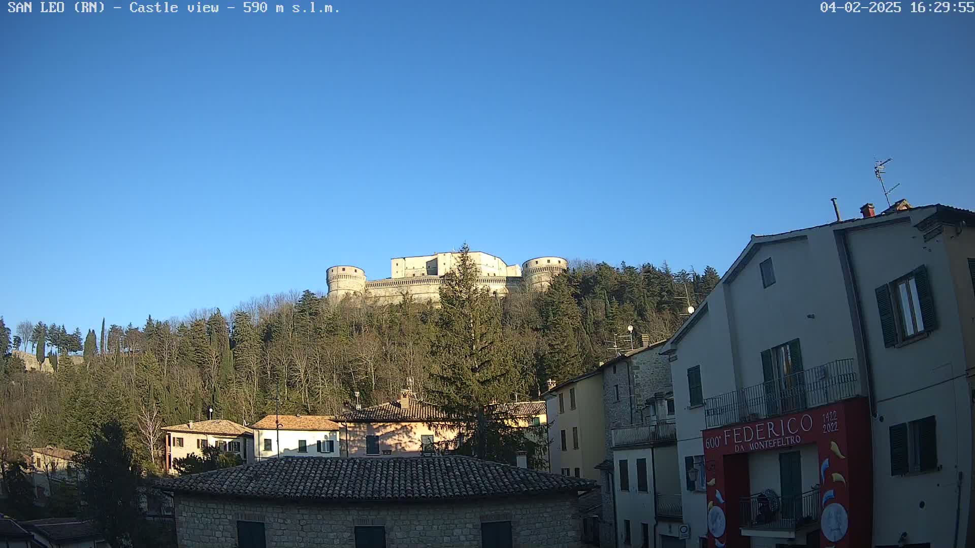 A large stone castle sits atop a tree-covered hill overlooking a town of stone buildings under a clear blue sky.