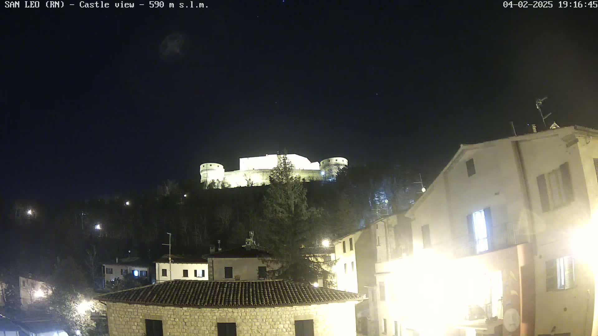 A brightly lit castle sits atop a hill overlooking a town at night under a clear, dark sky.