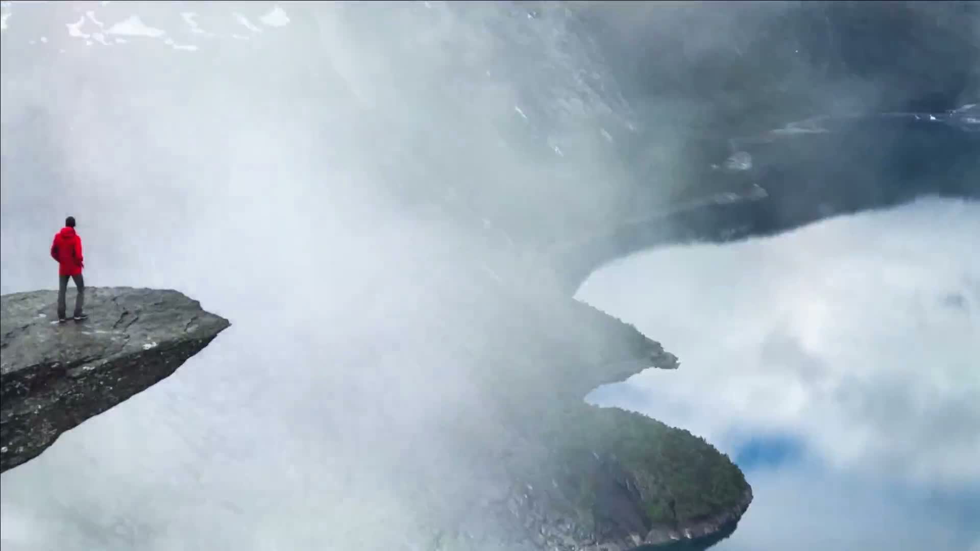 A person in a red jacket stands on a rocky precipice overlooking a foggy valley and a body of water.