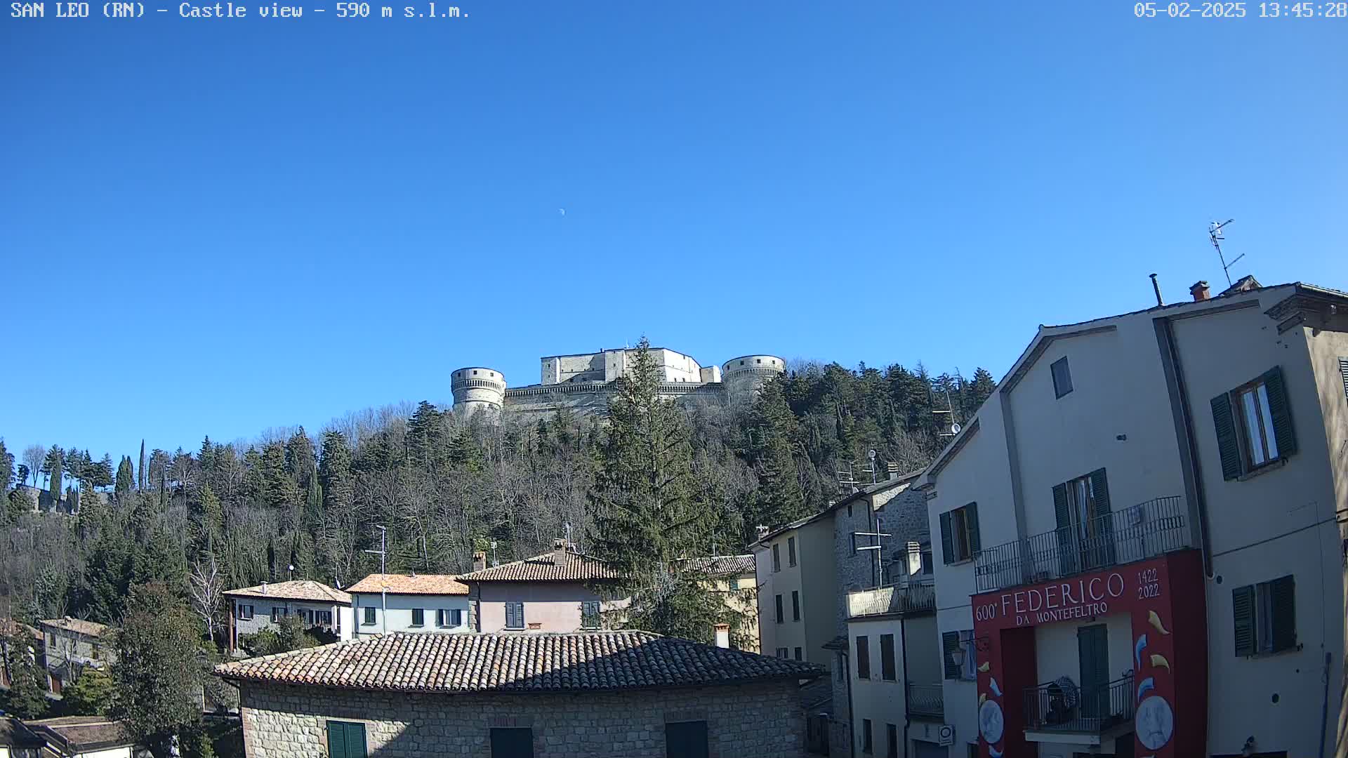 A stone castle sits atop a hill covered in leafless trees under a clear blue sky, overlooking a village of stone buildings.
