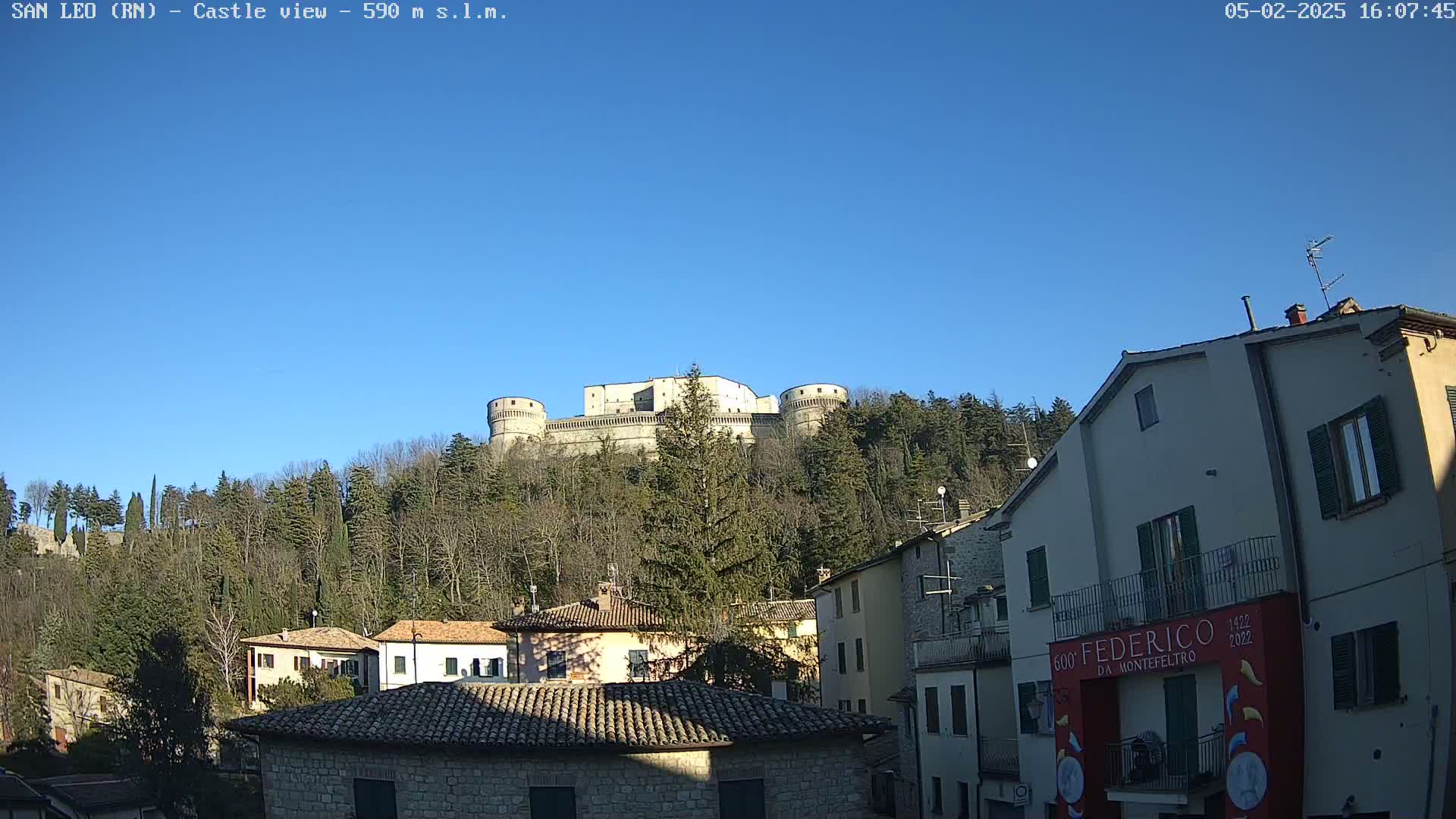 A stone castle sits atop a hill covered in leafless trees under a clear blue sky, overlooking a village of stone buildings.