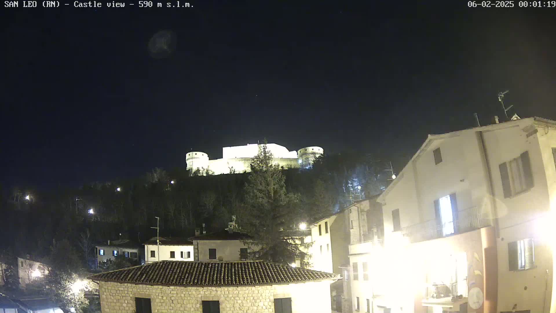 A brightly lit castle sits atop a hill at night, overlooking a town with buildings illuminated by streetlights under a clear, dark sky.